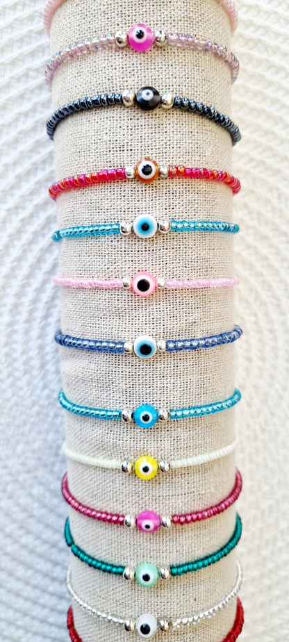 Pulsera elástica de protección ojo turco mini.  Realizada con mini bolas de rocalla en tonos brillantes, entre piezas de acero y detalle de ojo turco en distintos colores.